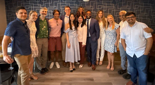 A group of eleven individuals pose in a contemporary indoor setting with blue geometric-patterned wallpaper and pendant lights. They are dressed in varied styles, ranging from professional attire like a blue suit and long dress to casual wear such as summer blouses and shorts. The overall atmosphere is cheerful and suggests a professional or semi-formal gathering.