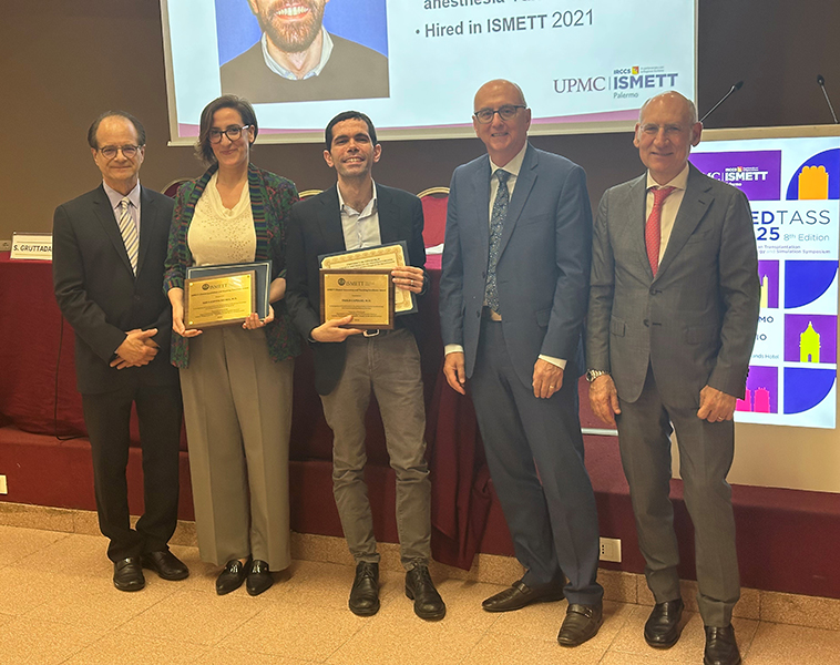Five individuals pose for a photo during the 8th MedTASS (Mediterranean Transplantation Anesthesiology and Simulation Symposium) 2025 event at ISMETT in Palermo, Italy. Two award recipients in the center hold plaques and certificates, while three other individuals, likely event organizers or faculty, stand on either side. Behind them, a screen displays a photo and text mentioning UPMC ISMETT and the MedTASS 2025 logo. The setting is a formal conference room with a red-carpeted stage and professional attire.
