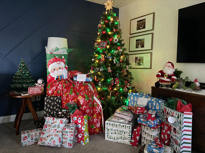 A festive room decorated for Christmas, featuring a lit Christmas tree adorned with colorful ornaments and lights. Surrounding the tree are numerous wrapped presents in various festive patterns, including Santa Claus and holiday-themed designs. A small table to the left holds a miniature Christmas tree and holiday decor, while a larger Santa Claus figure sits on a sideboard to the right. The walls have framed photos and a modern navy-blue accent design. The cozy atmosphere highlights a joyful holiday scene.