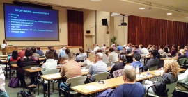 "A full classroom with people sitting at desks watching a lecture"