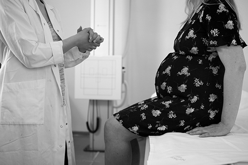 "A black and white photo of a doctor talking to a pregnant woman on a bed in a hospital room"