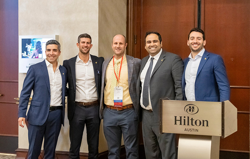 Five men in professional attire smile and pose together in a hallway at the Hilton Austin during the 2025 AAPM + MIT Hacking Medicine Innovation Challenge. The man in the center, Dr. Trent Emerick, wears a red lanyard with conference badges, indicating his participation. A podium with the Hilton Austin logo is visible to the right. The group appears to be part of a team or delegation, possibly presenters or award recipients at the event.