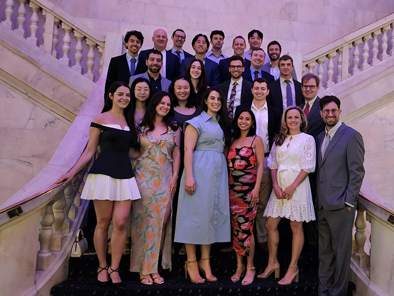 A group of 25 people dressed in formal and semi-formal attire pose for a group photo on a grand marble staircase with ornate railings. The group includes both men and women, smiling and standing in multiple rows, suggesting a celebratory or professional event in an elegant setting.