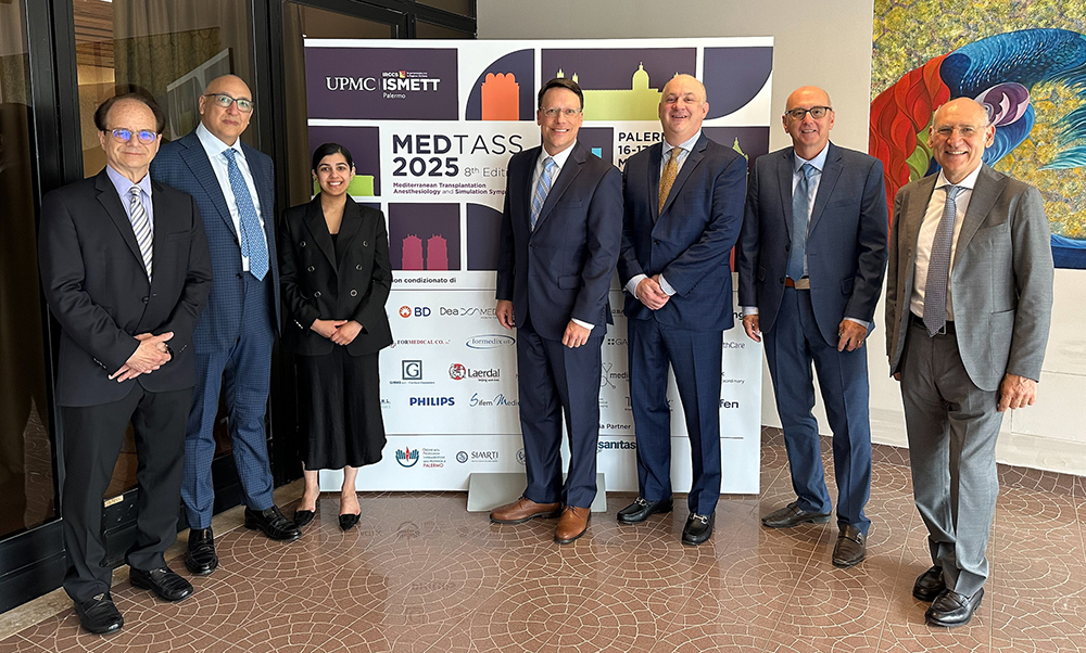 Seven professionally dressed individuals stand together in front of a banner for the 8th Edition of MEDTASS 2025 (Mediterranean Transplantation Anesthesiology and Simulation Symposium) held in Palermo. The banner features the UPMC and ISMETT logos, event dates (May 16–17, 2025), and logos of sponsoring organizations. The group poses in a hallway with tiled flooring and colorful artwork visible on the wall to the right.