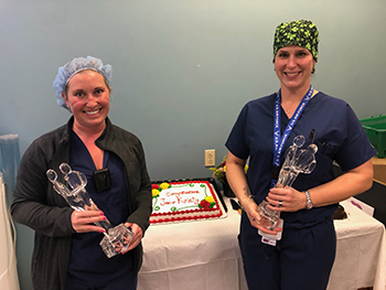 "Kristy and Jennifer pose with awards while in medical garb with a congratulatory cake in the background"