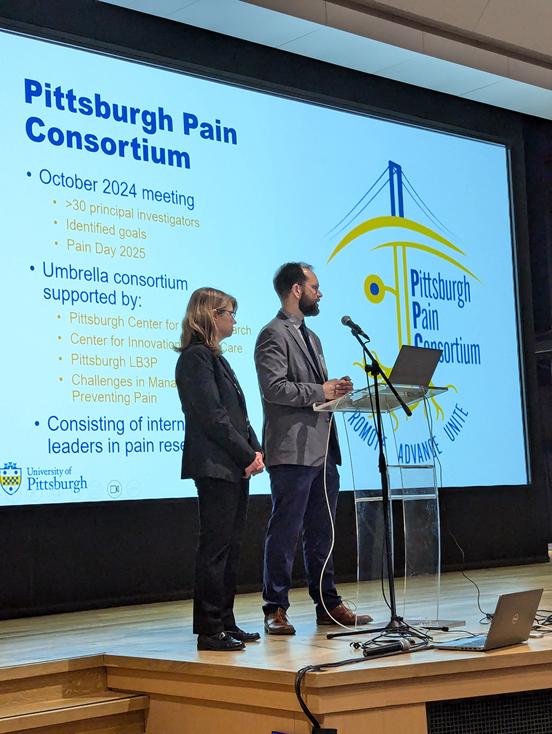 Two presenters stand at a podium on stage in front of a large screen displaying a presentation titled “Pittsburgh Pain Consortium.” The slide includes information about an October 2024 meeting, Pain Day 2025, and participating groups such as the Pittsburgh Center for Pain Research, Center for Innovation in Pain Care, and Pittsburgh LB3P. The Pittsburgh Pain Consortium logo, featuring a stylized bridge and neuron, is prominently displayed. The University of Pittsburgh logo appears at the bottom left of the screen. A laptop and microphone setup are visible on stage.