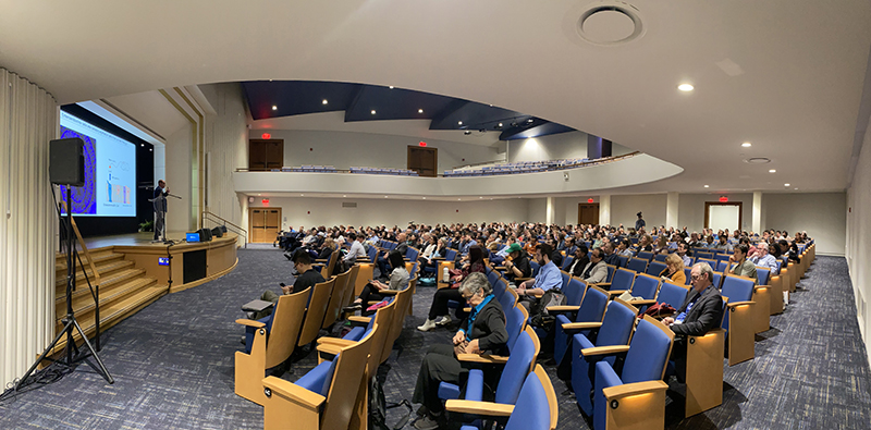 A wide-angle view of a large auditorium filled with attendees seated in rows of blue and wood chairs. Dr. David Julius presents on stage to the left, with a large projection screen showing a scientific slide. The room features a curved ceiling, balcony seating, and blue carpeting. The audience appears engaged, with many taking notes or listening attentively during the Pain Day 2025 event.