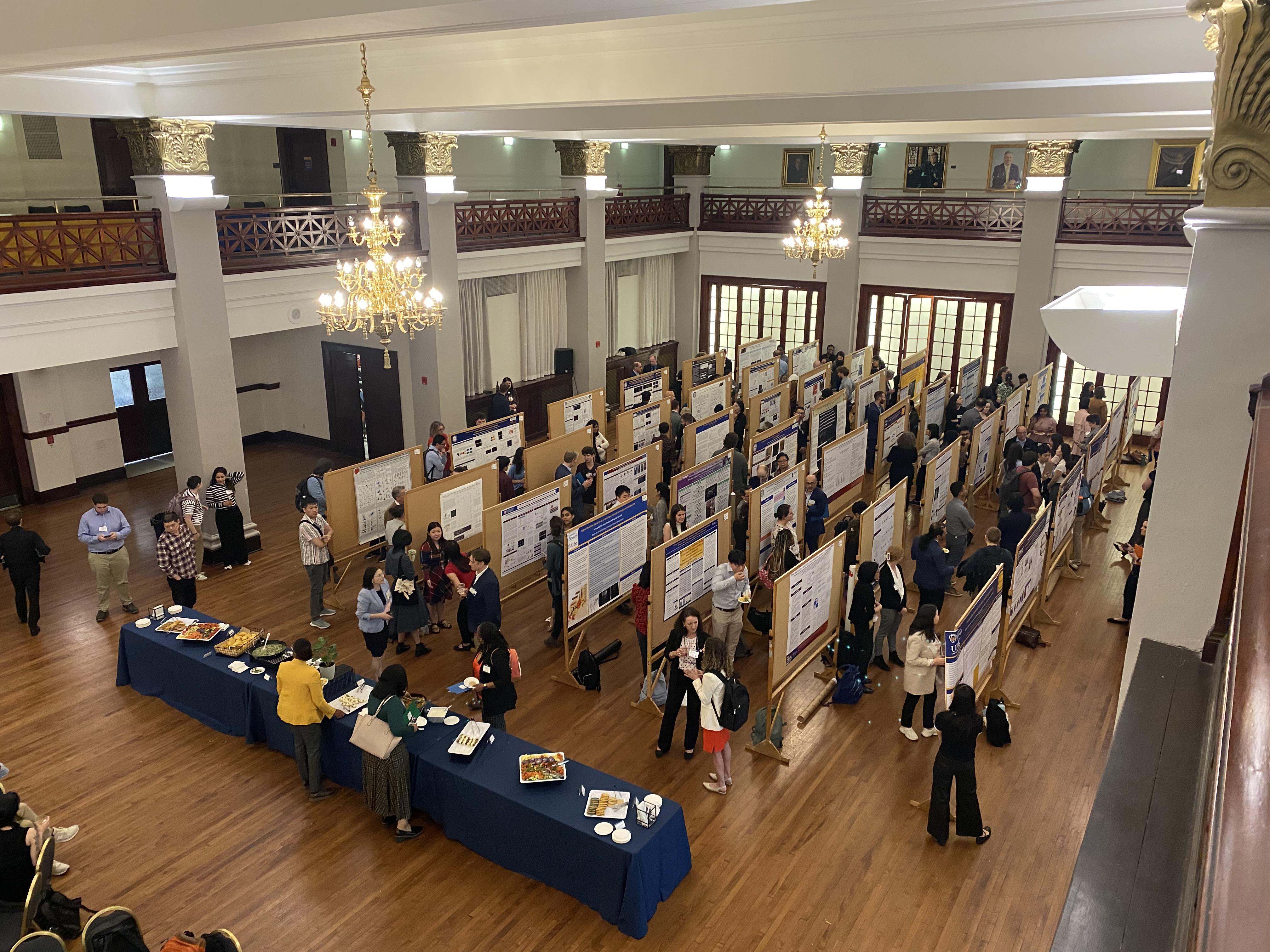 Overhead view of a bustling academic poster session held in a grand hall with ornate columns, chandeliers, and wooden floors. Rows of poster boards display research projects as attendees walk through, read posters, and engage in discussions. A long table with food and refreshments is set up along one side of the room. The event is part of Pain Day 2025, showcasing a vibrant exchange of ideas in a formal and elegant setting.