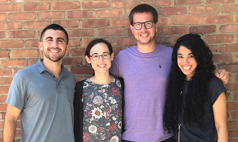 Four people stand close together smiling in front of a brick wall. The group includes two men and two women, all casually dressed. They appear relaxed and happy.