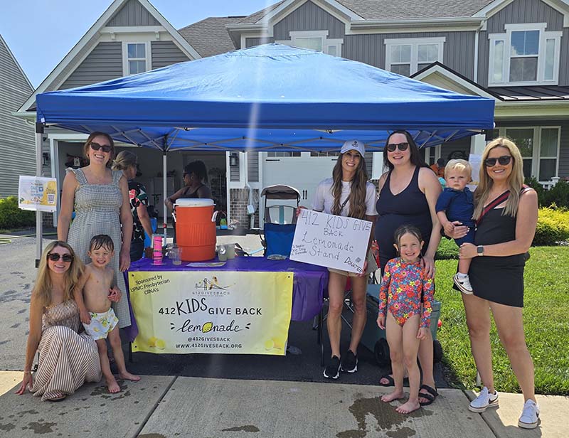 A lively neighborhood lemonade stand under a bright blue canopy on a sunny day. The stand is set up on a residential street lined with modern two-story houses. Underneath the canopy, a table with a vibrant yellow banner displaying "412 KIDS GIVE BACK" and a web address is visible. The table is filled with items likely supporting the lemonade sale, including a large orange cooler, purple pitchers, and cups. A homemade poster on the table reads "412 KIDS GIVE BACK Lemonade $1.00" with drawings of lemons, indicating the event's charitable purpose. Several children and adults are engaging with the stand. Two children are near the table, possibly waiting for a drink, while an adult behind the table serves or organizes items. Another adult with a baby in a carrier interacts across the table, and another adult holding a toddler stands at the table's far end. All individuals are dressed casually for a warm day, contributing to the event's casual, family-friendly atmosphere. The backdrop of well-maintained houses and clear skies suggests a community-oriented event focused on charity and family participation.