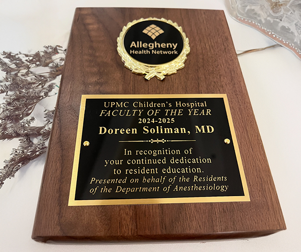 A wooden plaque with a gold and black plate awarded to Doreen Soliman, MD by UPMC Children's Hospital. The plaque reads: "FACULTY OF THE YEAR 2024–2025. In recognition of your continued dedication to resident education. Presented on behalf of the Residents of the Department of Anesthesiology." It also features the Allegheny Health Network logo at the top.