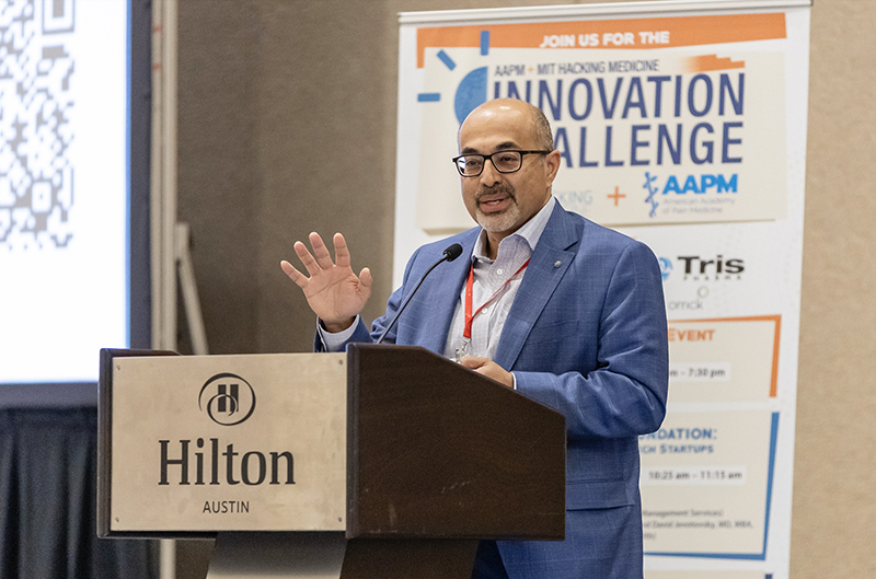 Dr. Ajay Wasan stands at a podium labeled "Hilton Austin," delivering a talk during the AAPM + MIT Hacking Medicine Innovation Challenge. He wears glasses, a blue suit, and a red lanyard, and gestures with one hand while speaking into the microphone. In the background, a vertical banner promotes the Innovation Challenge event, displaying logos for AAPM and MIT Hacking Medicine and a partial agenda. A large QR code is visible on the screen to the left.
