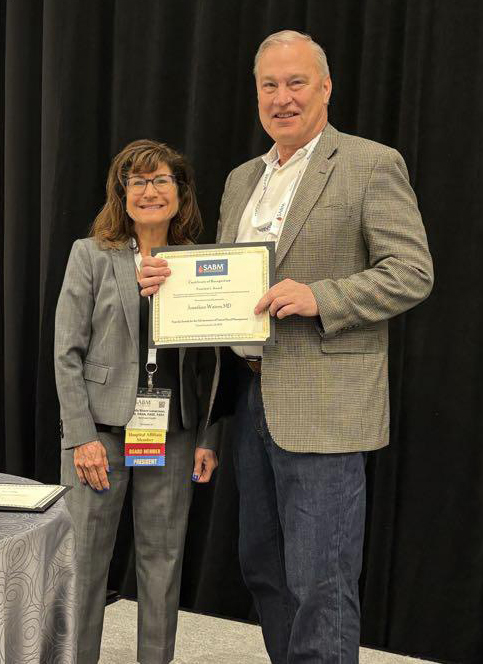A man and woman stand side by side smiling at an event. The man is holding an award certificate of from the Society for the Advancement of Blood Management (SABM), awarded to Jonathan Waters, MD. Both are dressed in business attire and wearing conference name badges. A black curtain serves as the background.