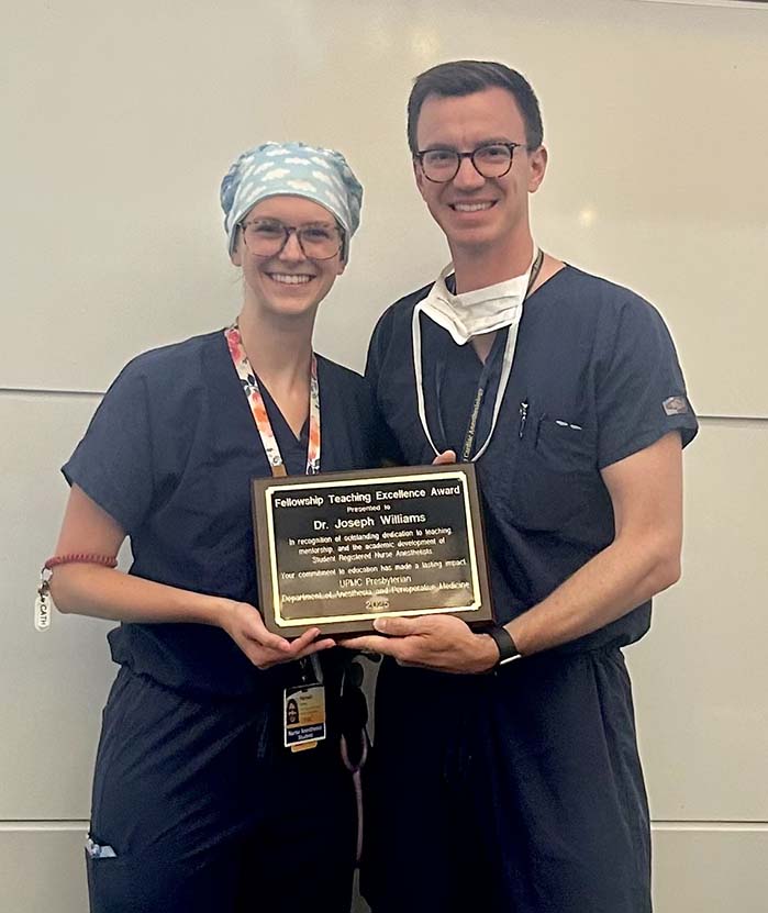 Two individuals dressed in medical attire stand together in a room, holding a plaque between them. The person on the left wears dark blue scrubs, an ID badge with an orange lanyard, and a light-colored surgical cap. The person on the right is in dark blue scrubs with a white undershirt, a stethoscope, and a face mask hanging below the chin. The plaque reads "Fellowship Teaching Excellence Award," presented to Dr. Joseph Williams for his dedication to teaching and mentorship.
