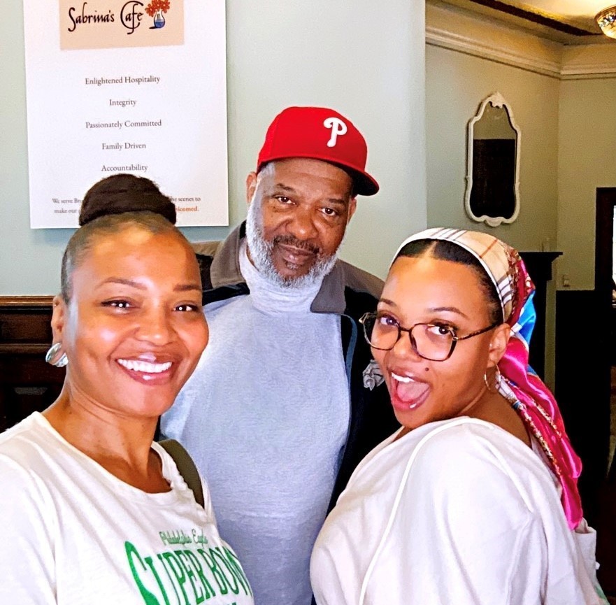 hree people smiling and posing together indoors at Sabrina's Café. The woman on the left wears a white T-shirt and has her hair in a high bun. The man in the center has a gray beard and wears a red Philadelphia Phillies cap and a gray sweatshirt. The woman on the right wears glasses, a colorful headscarf, and a white top, smiling with an open-mouthed expression. A sign with the café's values is visible on the wall behind them.