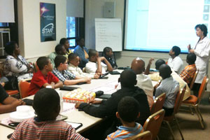 "A group of African American men and boys sitting around a table watching a lecture"