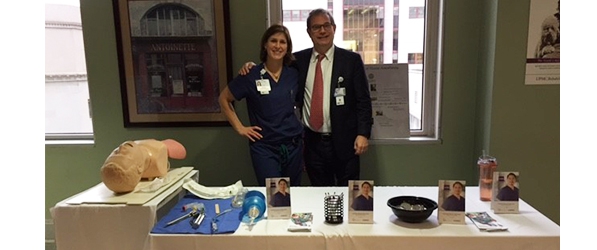 "Two people posing behind a table with medical instruments, flyers, and a training dummy"