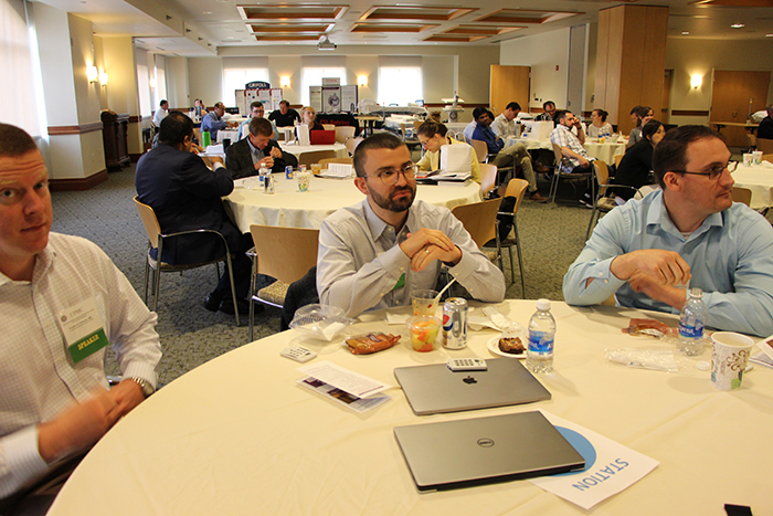 "Three men sit at a table in a large room with other people at tables"