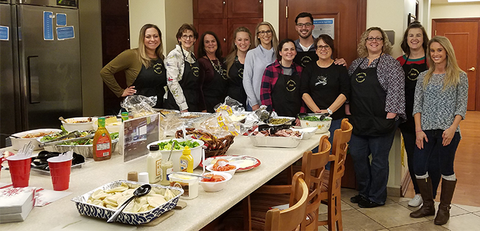 "A group of CRNAs posing in aprons in front of a table filled with food, expanded photo"