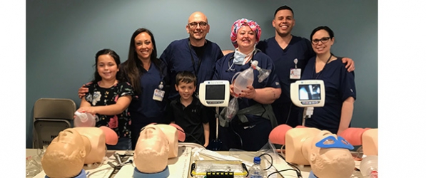 "A group of CRNAs and kids pose behind a table of dummies and medical equipment"