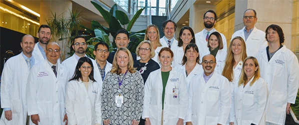 "A group of Clinicians, most of them in lab coats, pose for a group photo inside in front of some large plants"