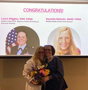 "Laura Wiggins and Danielle Meholic posing together with a bouquet of flowers with a congratulatory slide behind"