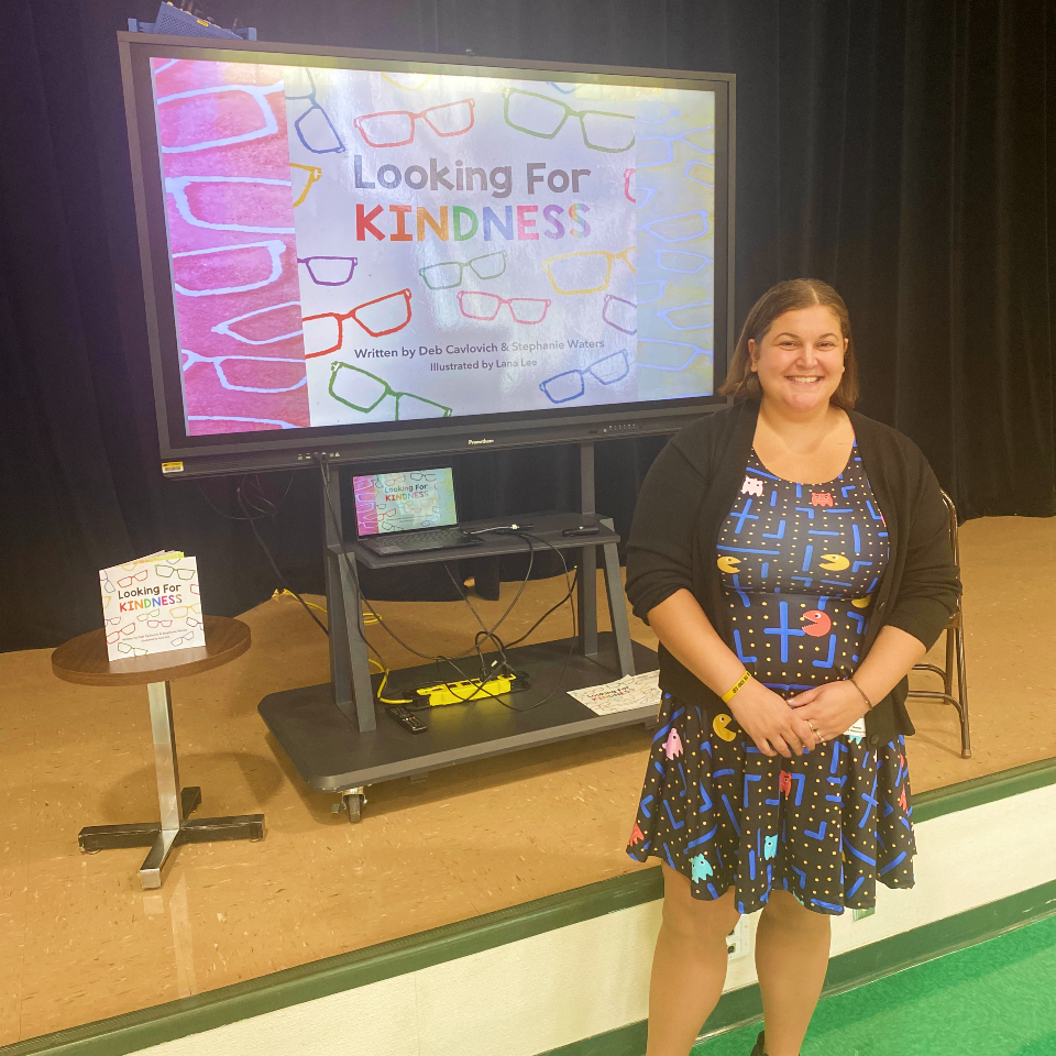 "Stephanie Waters poses in front of a screen with a slide reading Looking for Kindness"