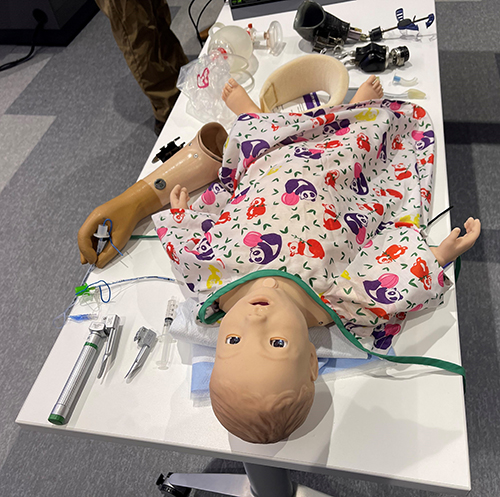 "A practice dummy of a child in a hospital gown on a table surrounded by medical instruments"