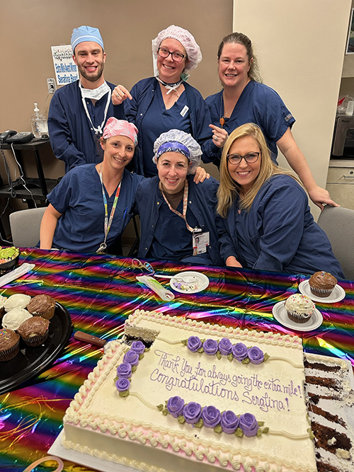 "A group photo of CRNAs in scrubs sitting behind a table with cupcakes and a congratulatory sheet cake"