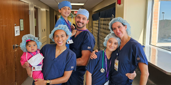 A group of six people, including adults and children, pose together in a hospital hallway. They are all wearing navy blue or pink scrubs and blue surgical caps, suggesting a medical or healthcare setting. One woman holds a baby dressed in pink scrubs with a badge that reads "Future CRNA." Everyone is smiling, and the atmosphere is warm and cheerful. A large window on the right lets in natural light.