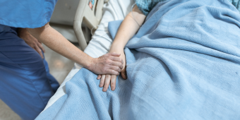 A close-up image of a healthcare professional gently holding the hand of a patient lying in a hospital bed. The patient is covered with a light blue blanket and wearing a hospital gown. The caring gesture conveys comfort, support, and compassion in a clinical setting.