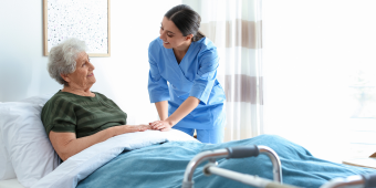 A smiling nurse in blue scrubs leans over to speak with an elderly female patient sitting upright in a hospital bed. The patient, wearing a green top, looks back at the nurse with a smile. A walker is visible in the foreground, and the room is bright with natural light coming through sheer curtains. The interaction conveys warmth, reassurance, and attentive care in a clinical setting.