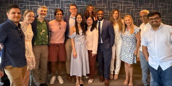 A group of eleven individuals pose in a contemporary indoor setting with blue geometric-patterned wallpaper and pendant lights. They are dressed in varied styles, ranging from professional attire like a blue suit and long dress to casual wear such as summer blouses and shorts. The overall atmosphere is cheerful and suggests a professional or semi-formal gathering.
