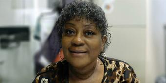 A woman with short, curly black hair is smiling at the camera. She is wearing large gold hoop earrings and a black and brown leopard-print top with a brooch on the left side. The background includes an office setting with a filing cabinet. A circular sticker with a colorful design is visible on the cabinet.
