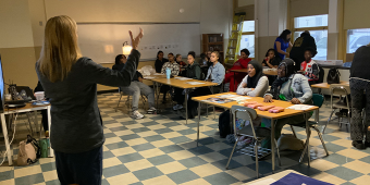 A woman stands at the front of a classroom speaking to a group of attentive high school students seated at desks. The students are watching and listening as she gestures with her hand. The classroom has checkered tile floors, large windows, and educational materials on the desks. A few other adults are visible in the background, assisting or observing.