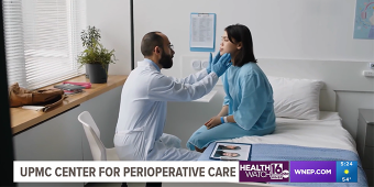 A doctor wearing a white coat and gloves examines a young woman in a hospital gown who is sitting on a bed in a medical setting. A clipboard with documents and medical images is on the bed. The room has a window with natural light, a potted plant, and a desk with a bag and papers. The banner at the bottom reads "UPMC Center for Perioperative Care," with a "Health Watch 16" and "WNEP.com" logo, and the time and temperature displayed as 5:24 and 54°F.