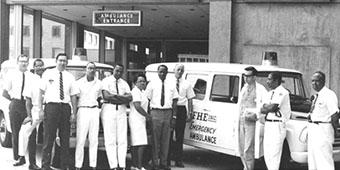 A black and white photo of a group of people standing in front of an ambulance in the year 1968