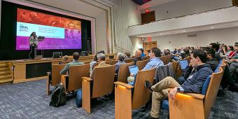 A speaker presents on stage in a large auditorium during a Pain Day 2025 session. The presentation slide reads “Back Pain Consortium (BACPAC) Research Program Overview.”. The audience, seated in wooden theater chairs, listens attentively, with some attendees taking notes on laptops. The room features a high ceiling, blue carpet, and a mix of lower and upper seating areas. A “HEAL Initiative” logo appears on the screen alongside vibrant background images.