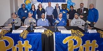 A group photo at the University of Pittsburgh first signing day for startups event featuring 13 people, including Dr. Trent Emerick, standing or seated behind tables adorned with blue and gold Pitt banners and pom-poms. The group includes individuals in Pitt apparel, the Pitt mascot Roc the Panther, and others dressed in business or casual attire. The backdrop includes balloons and a Pitt logo.