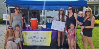 A lively neighborhood lemonade stand under a bright blue canopy on a sunny day. The stand is set up on a residential street lined with modern two-story houses. Underneath the canopy, a table with a vibrant yellow banner displaying "412 KIDS GIVE BACK" and a web address is visible. The table is filled with items likely supporting the lemonade sale, including a large orange cooler, purple pitchers, and cups. A homemade poster on the table reads "412 KIDS GIVE BACK Lemonade $1.00" with drawings of lemons, indi