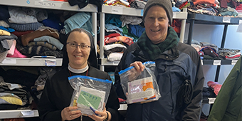 A nun and a man stand in a room filled with shelves stacked with folded clothing. They are both smiling and holding clear plastic bags containing care package items, such as snacks and hygiene products. The nun’s bag has a handwritten note that reads “HAPPY.” In front of them is a large cardboard box labeled “Cara Cara Oranges,” filled with more care packages. The setting appears to be a donation or volunteer center, where supplies are being prepared for distribution.