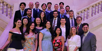 A group of 25 people dressed in formal and semi-formal attire pose for a group photo on a grand marble staircase with ornate railings. The group includes both men and women, smiling and standing in multiple rows, suggesting a celebratory or professional event in an elegant setting.