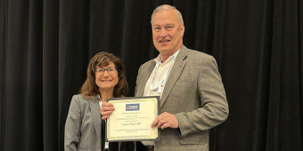 A man and woman stand side by side smiling at an event. The man is holding an award certificate of from the Society for the Advancement of Blood Management (SABM), awarded to Jonathan Waters, MD. Both are dressed in business attire and wearing conference name badges. A black curtain serves as the background.