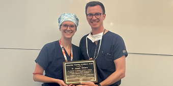 Two individuals dressed in medical attire stand together in a room, holding a plaque between them. The person on the left wears dark blue scrubs, an ID badge with an orange lanyard, and a light-colored surgical cap. The person on the right is in dark blue scrubs with a white undershirt, a stethoscope, and a face mask hanging below the chin. The plaque reads "Fellowship Teaching Excellence Award," presented to Dr. Joseph Williams for his dedication to teaching and mentorship.